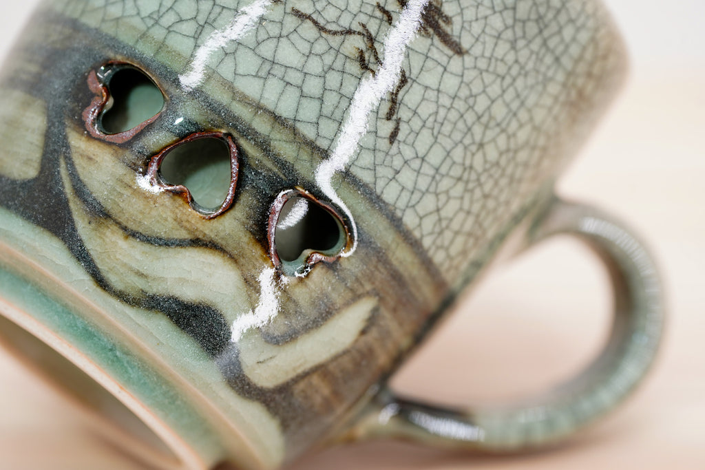 Angled close-up of the Soma mug’s heart-shaped openings, swirling brown-green glaze and curved handle.