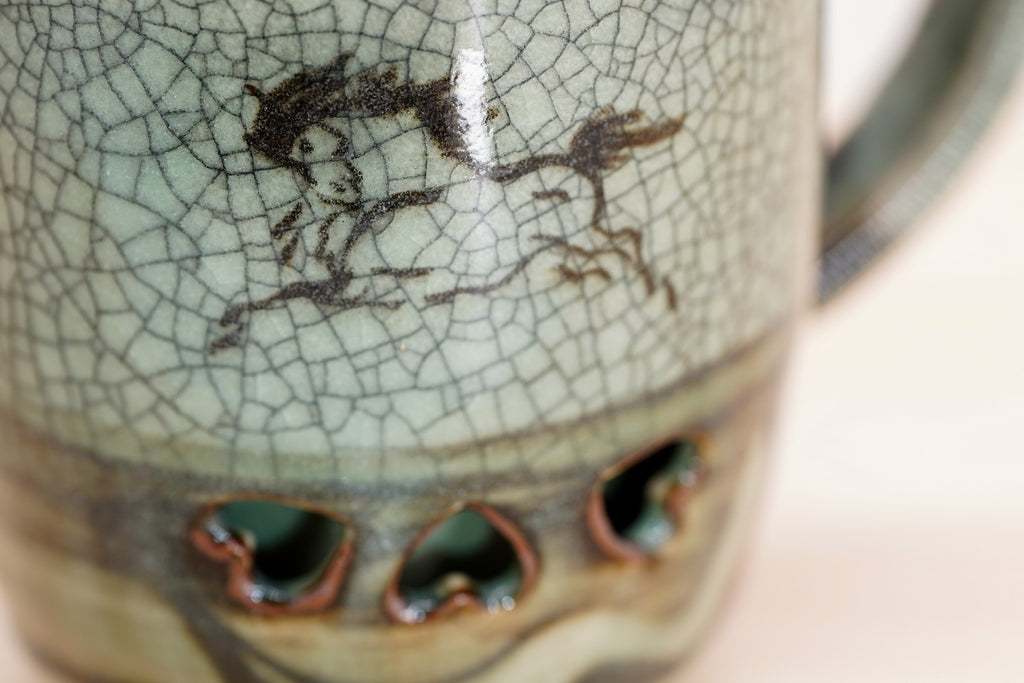 Close-up of the Soma mug’s running horse on grey-green crackle glaze above the dark lower band.